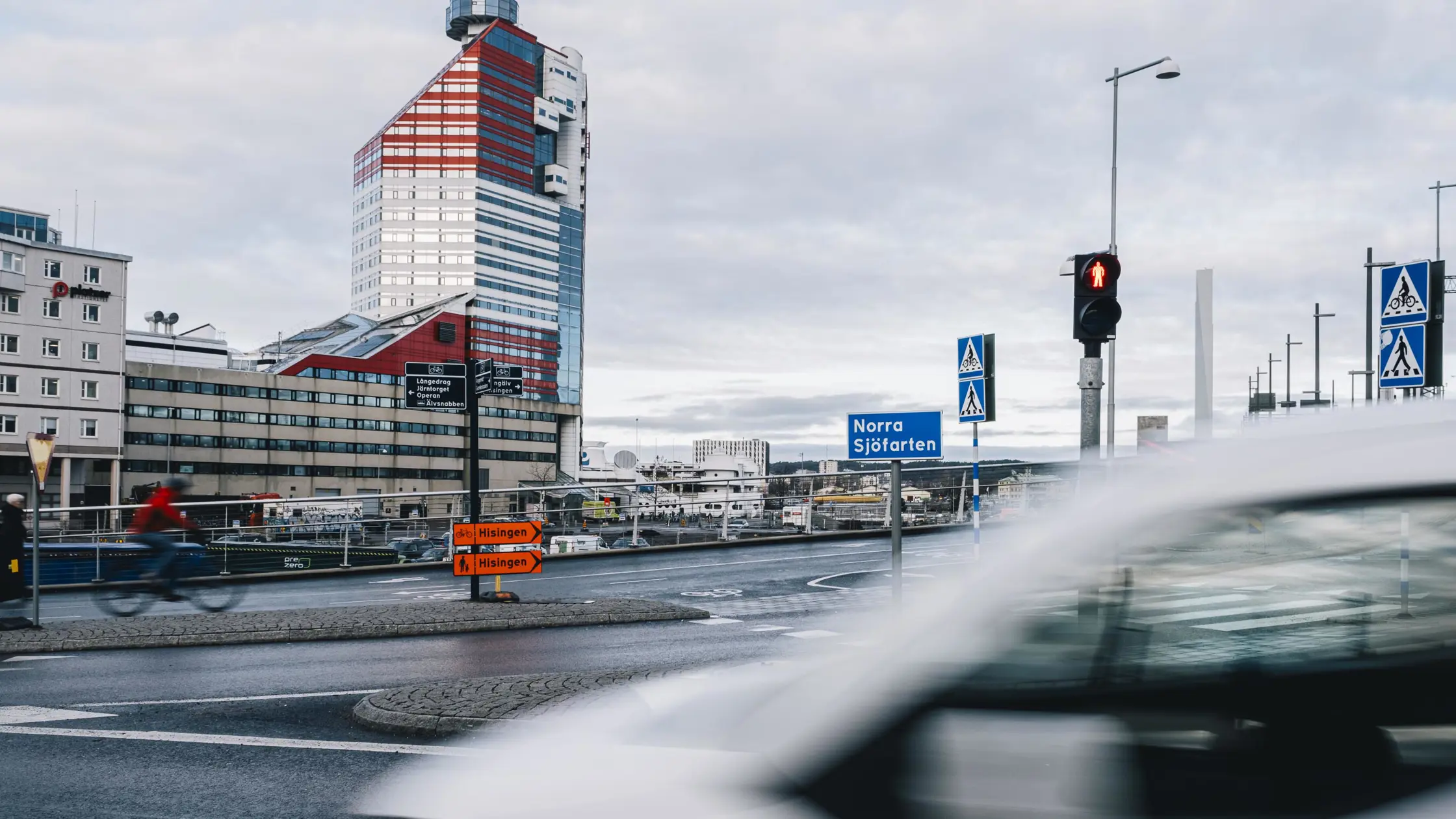 En vit bil och en cyklist susar förbi på Norra Sjöfarten med huset kallat Läppstiftet i bakgrunden.