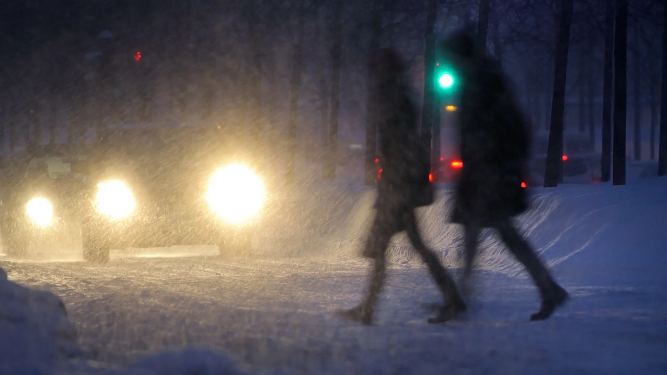 Bilden visar två mörka gestalter som tar sig över en snöigväg i billampornas sken utan reflex.